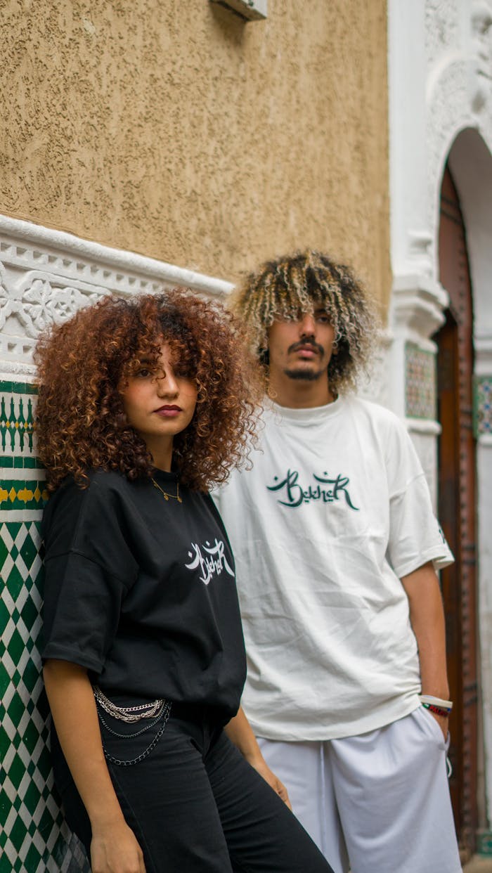 service-01 Young stylish couple posing in urban streetwear, leaning against decorative wall outdoors.
